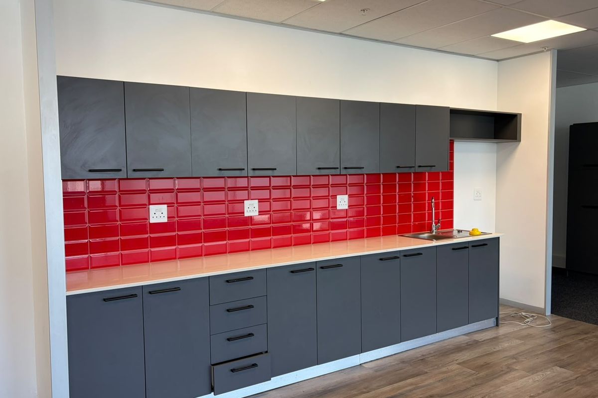 A modern office kitchenette featuring dark grey cabinetry, a vibrant red subway tile backsplash, and a stainless steel sink.