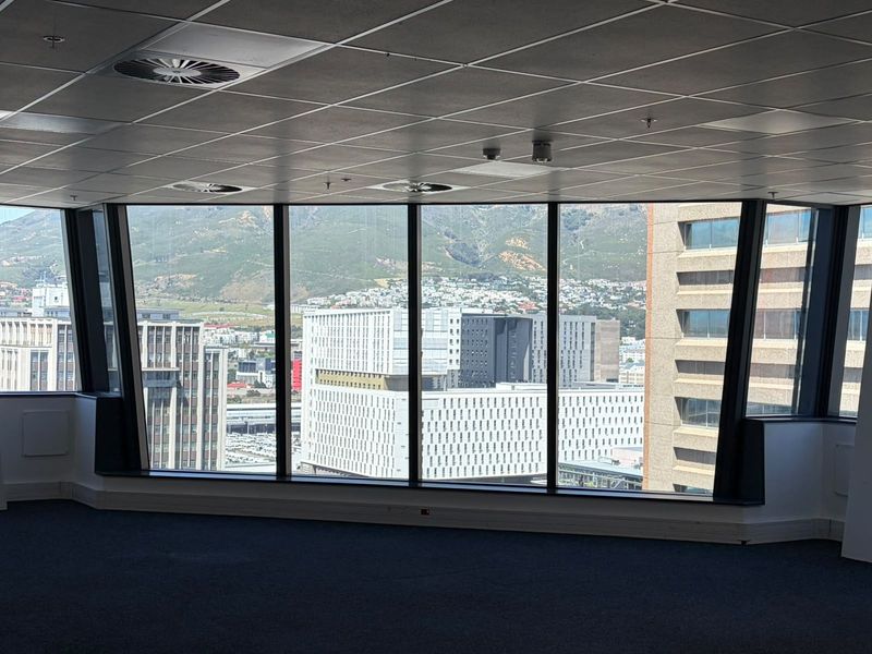 A spacious, empty office suite featuring expansive floor-to-ceiling windows with panoramic city and mountain views, suspended ceilings, and carpeted flooring.