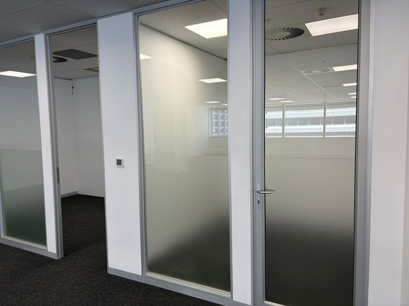 Modern office space featuring glass partition walls with frosted privacy panels, suspended ceilings with recessed lighting, and clean carpet flooring.