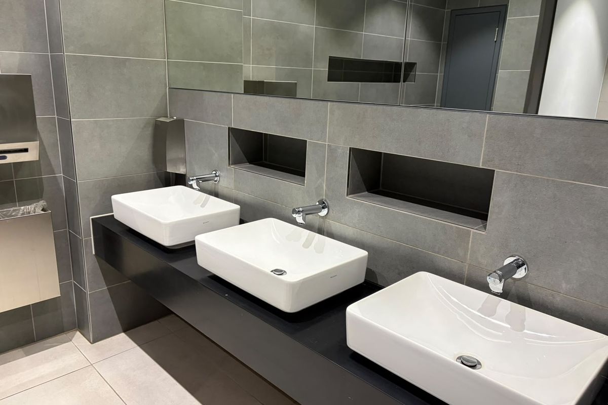 Modern commercial bathroom featuring a sleek triple vessel sink vanity with wall-mounted faucets, large mirror, and contemporary grey tiled walls.