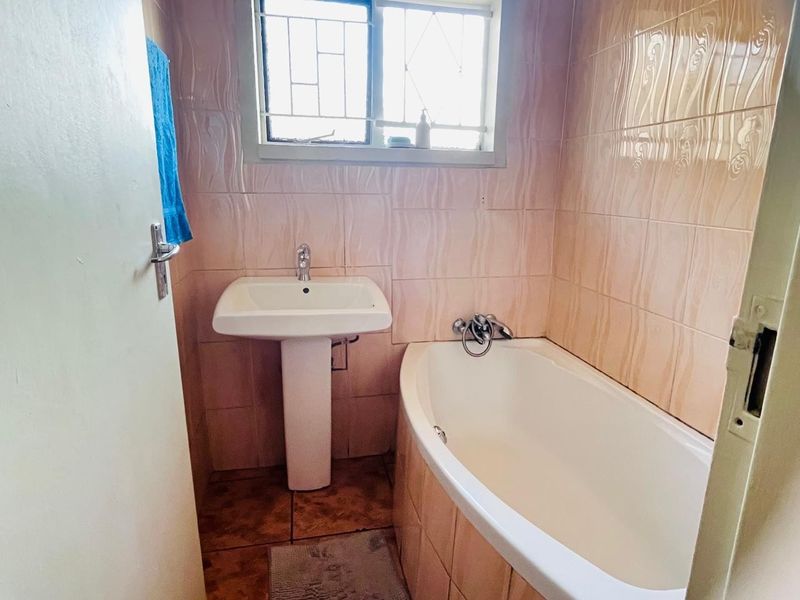 A compact bathroom featuring a pedestal washbasin, a built-in bathtub, and full-height wall tiling with security bars on the window.