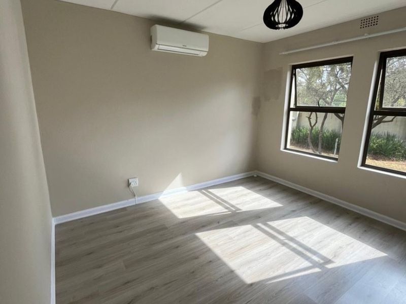 A bright, empty bedroom featuring modern laminate flooring, an air conditioning unit, and large windows with natural light.