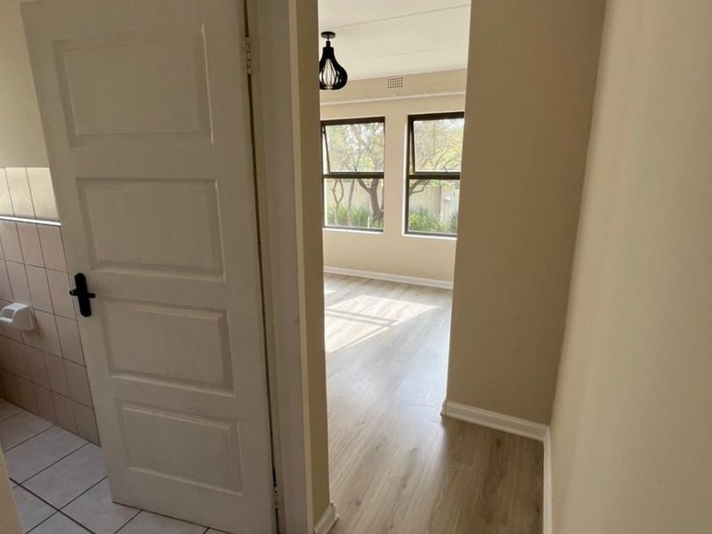 A bright hallway featuring neutral wall tones, laminate flooring, and a tiled transition area leading into an adjacent room with large windows.