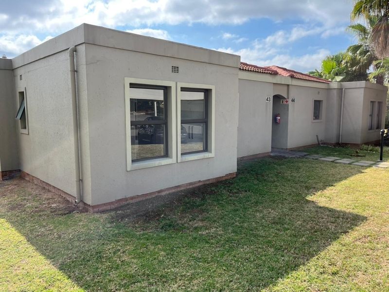 Front elevation of a single-story residential unit featuring a plastered facade, tiled roof, and aluminium window frames with an adjacent lawn area.