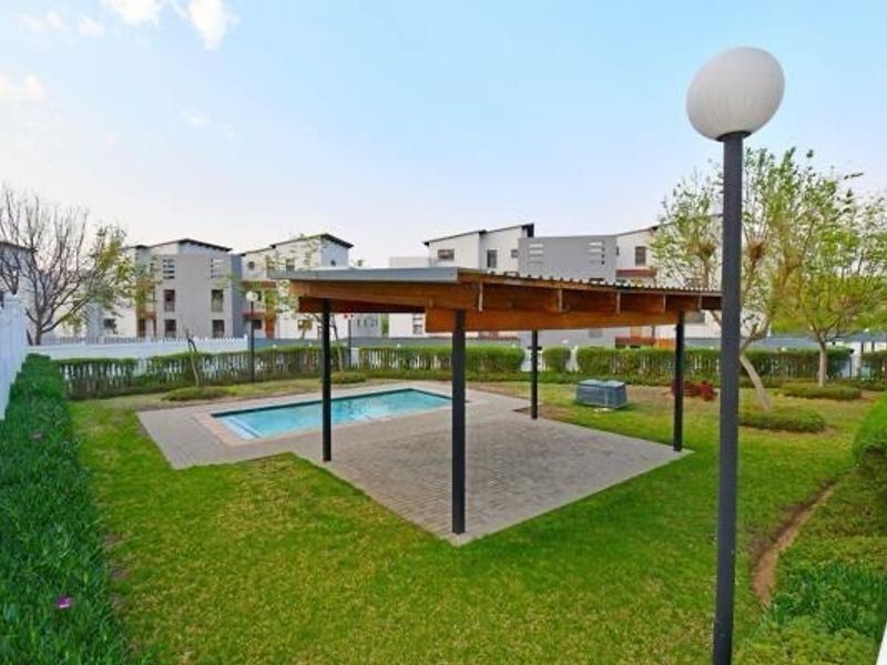 Communal outdoor area featuring a swimming pool, a covered patio structure with paved flooring, and landscaped garden surroundings.