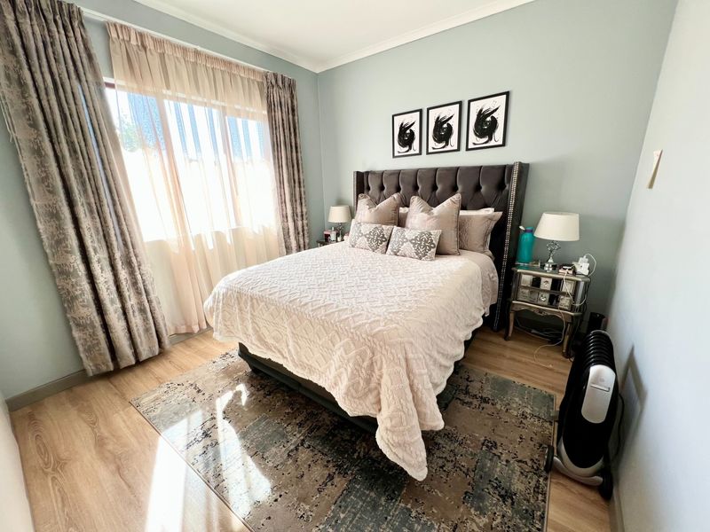 A well-maintained bedroom featuring light-toned laminate flooring, soft blue wall paint, and a large window with floor-to-ceiling curtains.