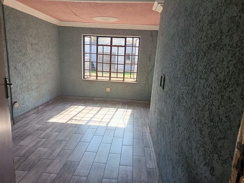 An empty living room featuring textured walls, wood-look floor tiles, and a ceiling rose, requiring minor repairs to the ceiling and electrical fixtures.
