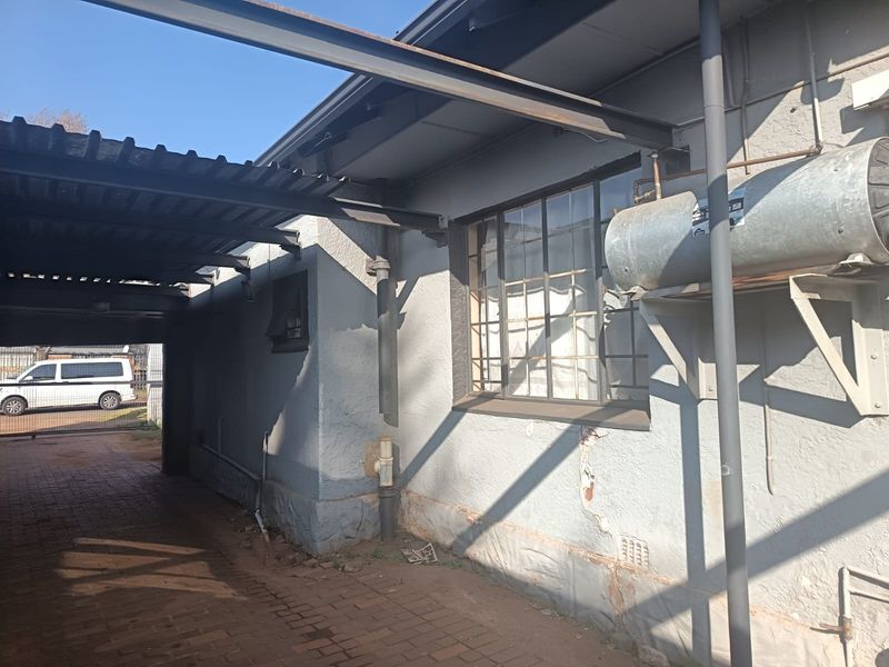 Side exterior view showing a carport, brick paving, and an external geyser unit mounted on a wall with visible plaster damage.