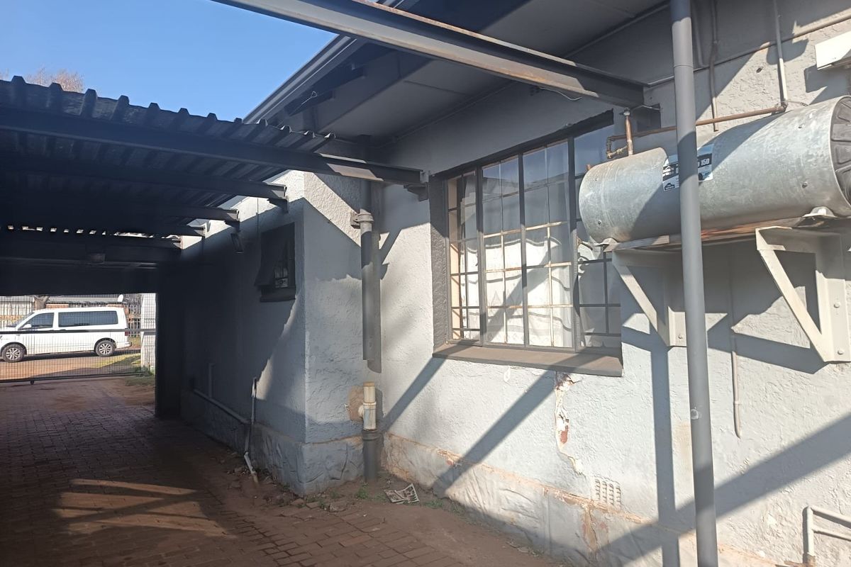 Side exterior view showing a carport, brick paving, and an external geyser unit mounted on a wall with visible plaster damage.
