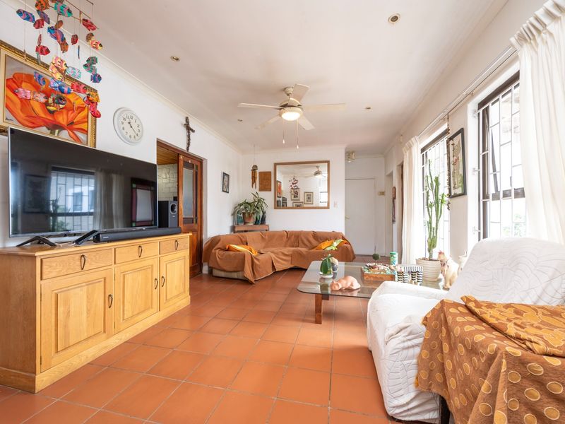 A spacious living room featuring terracotta floor tiles, a ceiling fan, and large windows with security bars, offering a comfortable and functional living space.