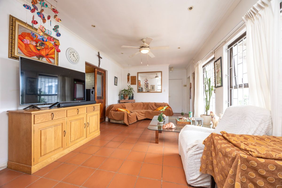 A spacious living room featuring terracotta floor tiles, a ceiling fan, and large windows with security bars, offering a comfortable and functional living space.