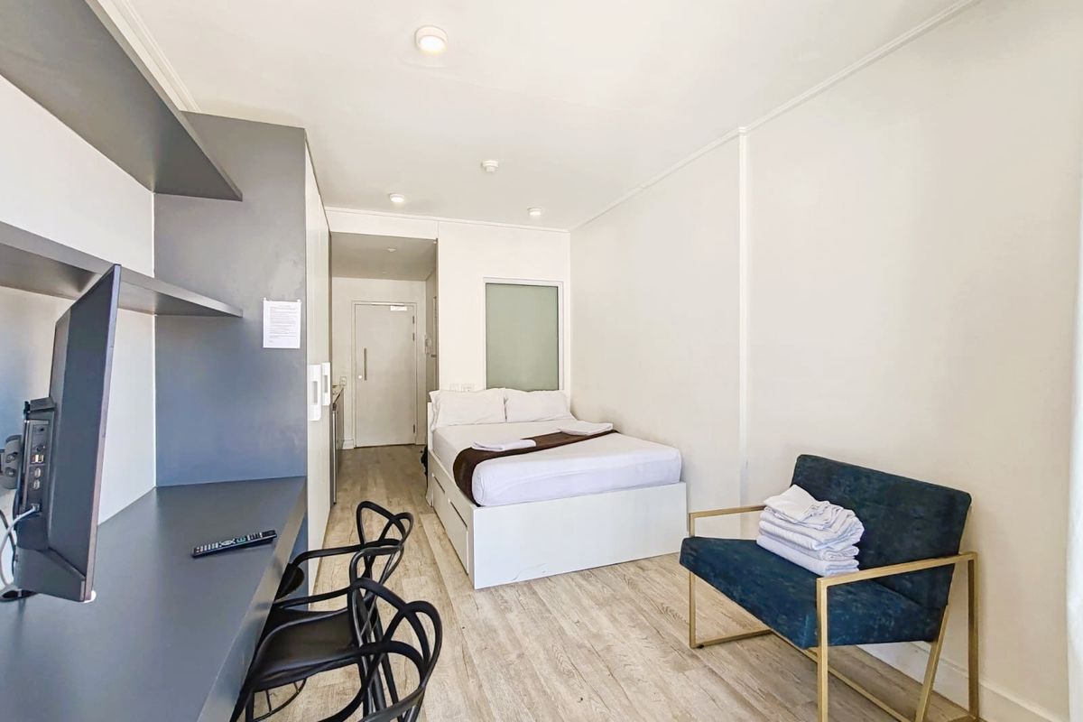 A compact studio bedroom featuring light wood-look laminate flooring, built-in desk and shelving unit, and recessed ceiling lighting.
