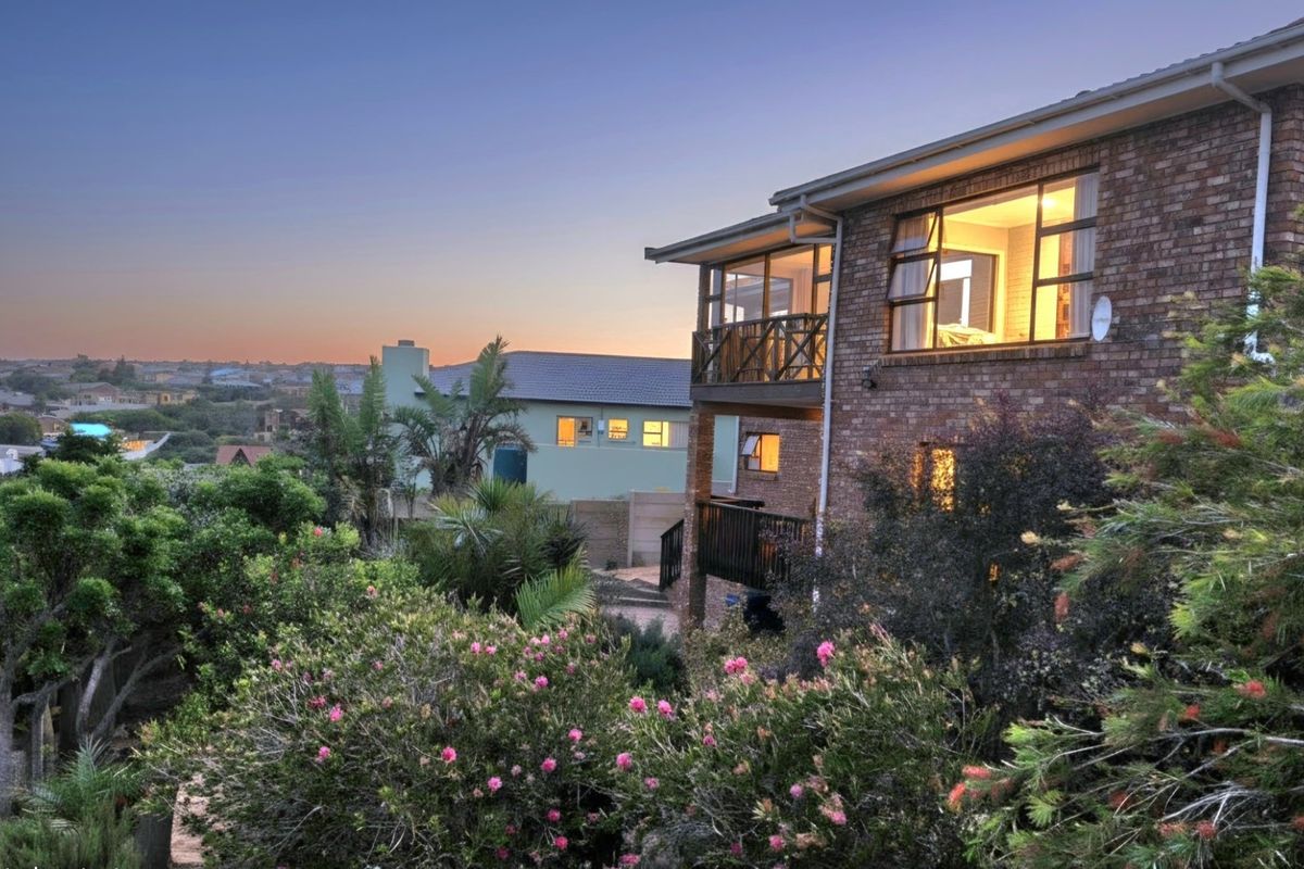 A rear view of a multi-story brick house at dusk, featuring a balcony with timber balustrades and established garden landscaping.