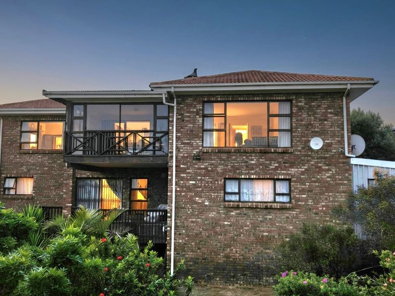 Rear elevation of a double-storey face brick house featuring a balcony, tiled roof, and established garden surroundings at dusk.