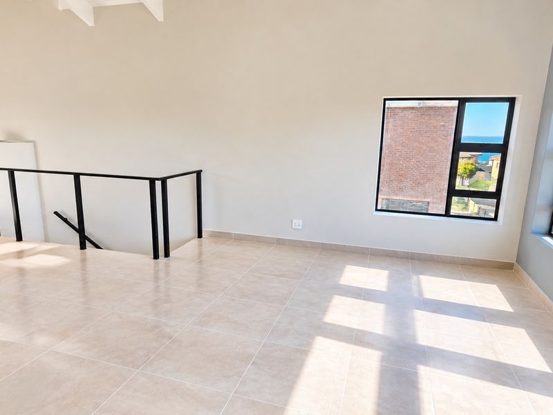 A bright, open-plan living area featuring neutral tiled flooring, exposed ceiling beams, and large windows with coastal views.