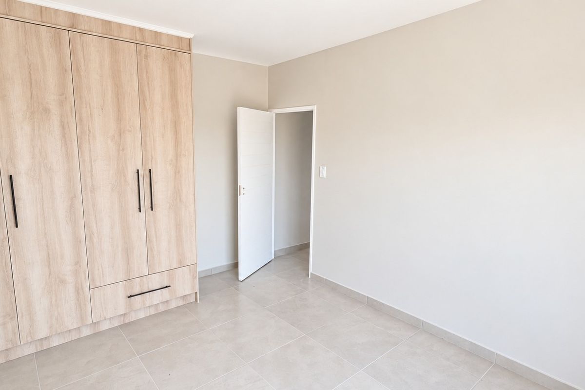 A bright, empty bedroom featuring modern light wood-finish built-in cupboards and neutral tiled flooring.