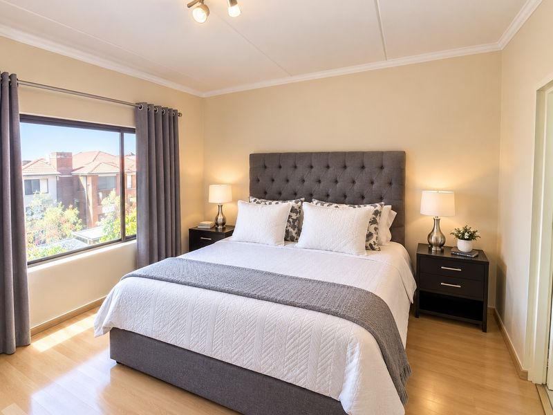 A well-maintained bedroom featuring light laminate flooring, neutral wall tones, and a large window providing natural light and a view of the surrounding townhouse complex.