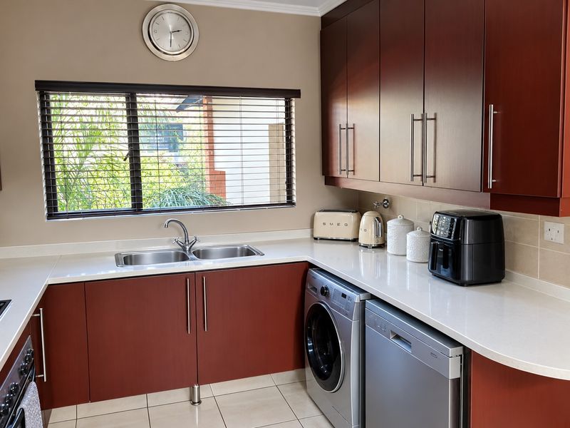 A well-maintained kitchen featuring dark wood cabinetry, quartz countertops, a double bowl stainless steel sink, and tiled flooring with a neutral backsplash.