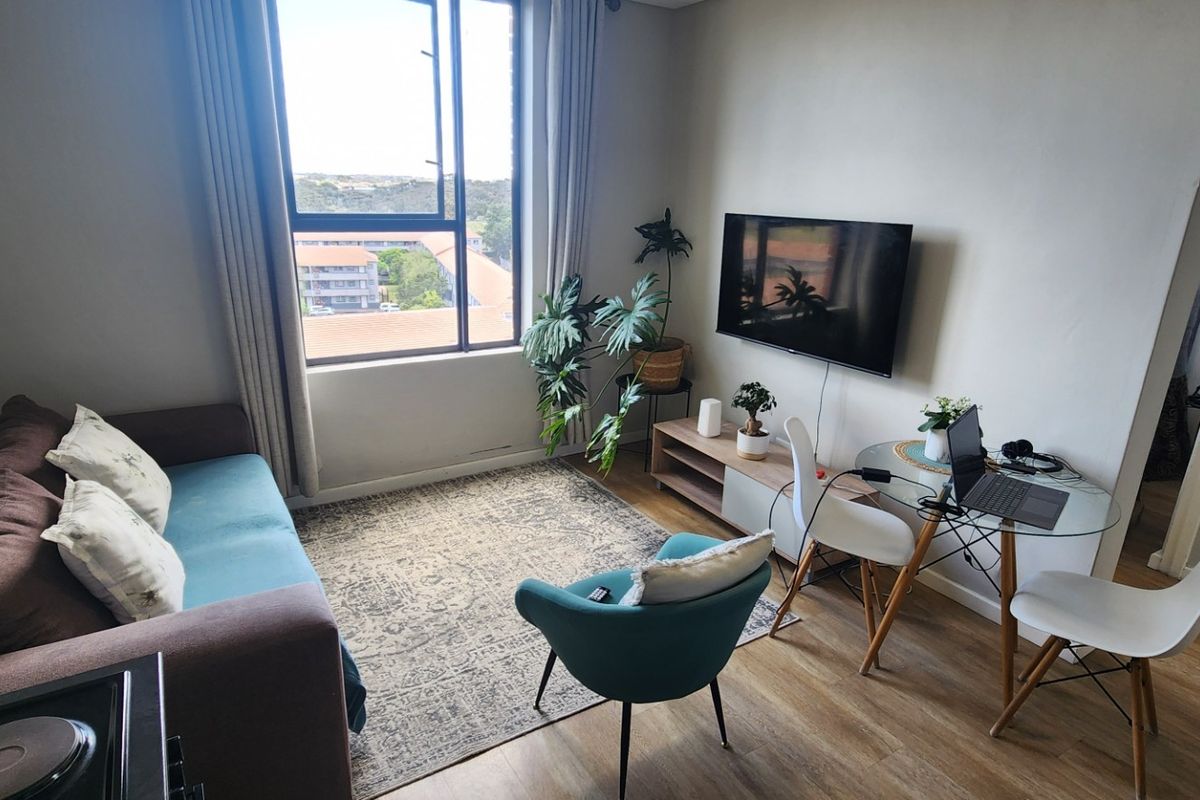 A bright living room featuring laminate flooring, a large window with natural light, and a wall-mounted television.