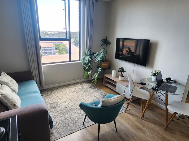 A bright living room featuring laminate flooring, a large window with natural light, and a wall-mounted television.