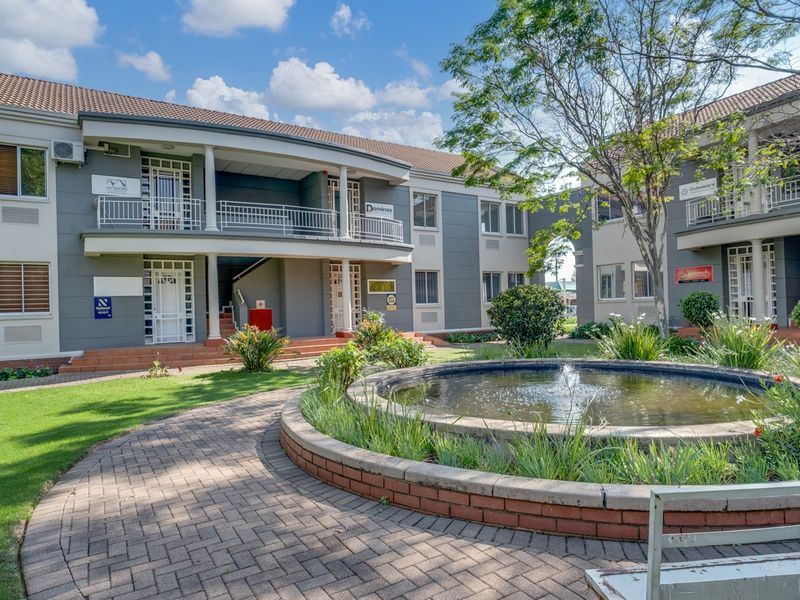 A well-maintained office complex exterior featuring a landscaped courtyard with a central water fountain, paved walkways, and multi-level office units with balconies.