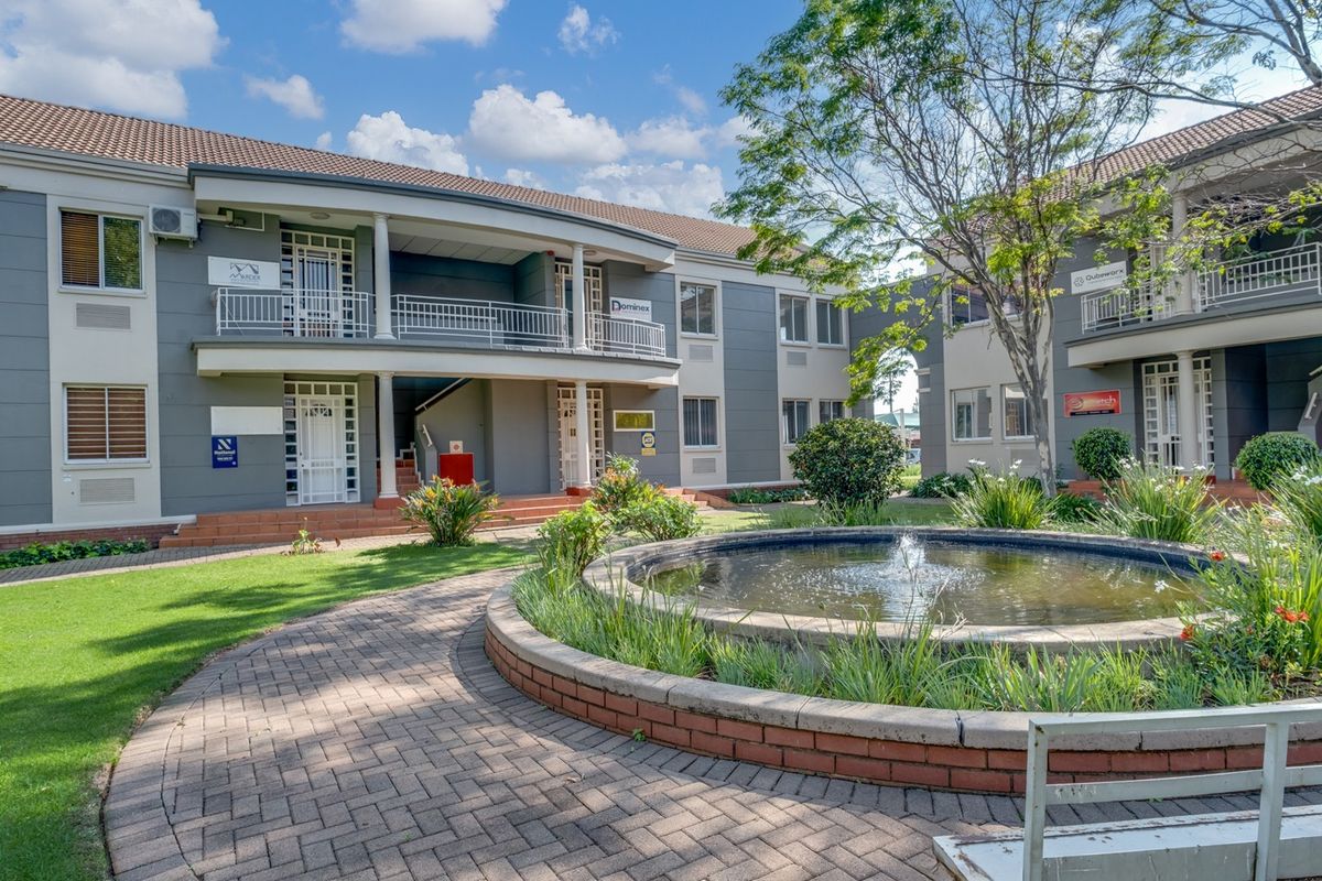 A well-maintained office complex exterior featuring a landscaped courtyard with a central water fountain, paved walkways, and multi-level office units with balconies.