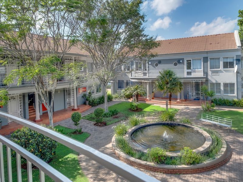 A well-maintained office complex courtyard featuring a central water fountain, paved walkways, landscaped gardens, and multi-level building access with balconies.