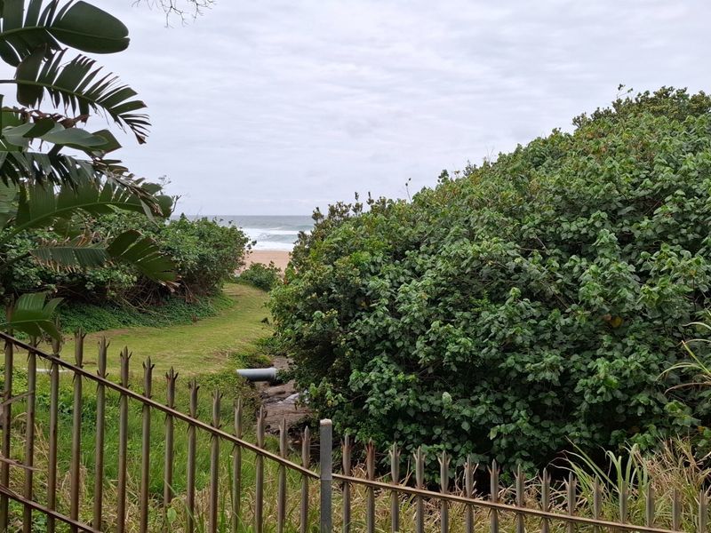 A view of the coastal landscape and beach access path, framed by established vegetation and metal perimeter fencing.
