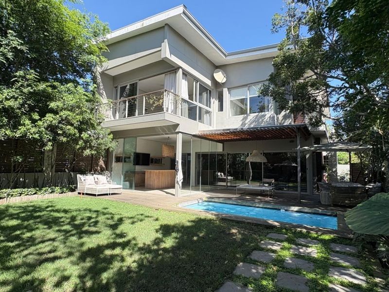 Rear view of a modern townhouse featuring a swimming pool, wooden decking, covered patio, and an upper-level balcony overlooking a landscaped garden.