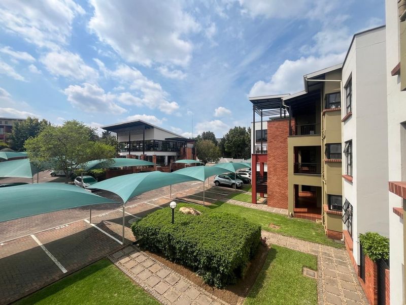 View of the apartment complex exterior featuring landscaped gardens, paved parking areas with shade cloth carports, and multi-story brick and plaster residential buildings.
