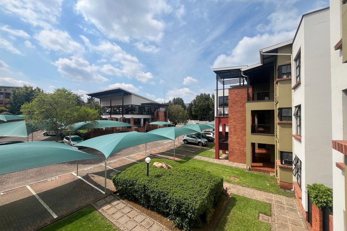 View of the apartment complex exterior featuring landscaped gardens, paved parking areas with shade cloth carports, and multi-story brick and plaster residential buildings.