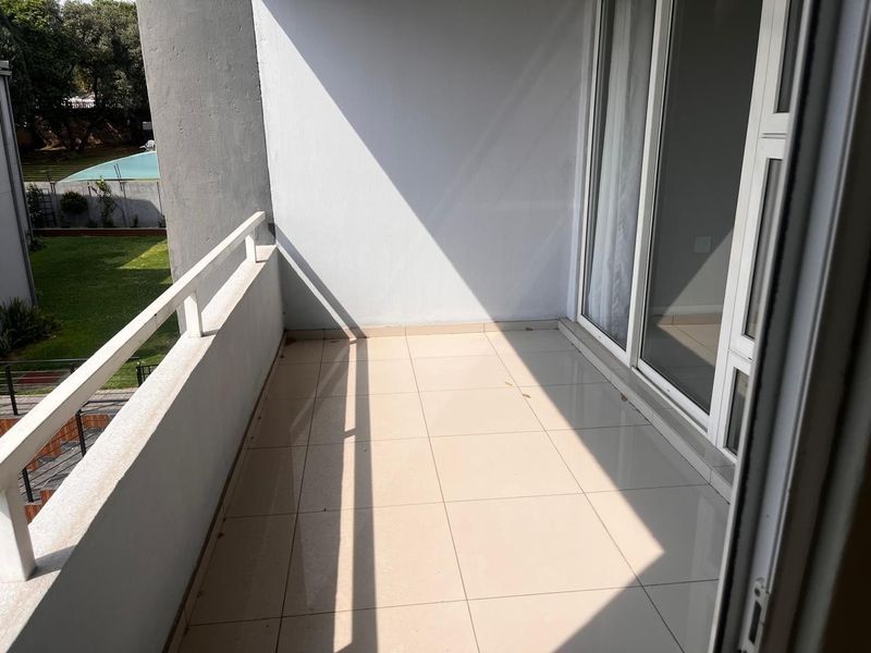 A clean, tiled balcony featuring a metal balustrade and sliding glass door access, overlooking a communal garden area.