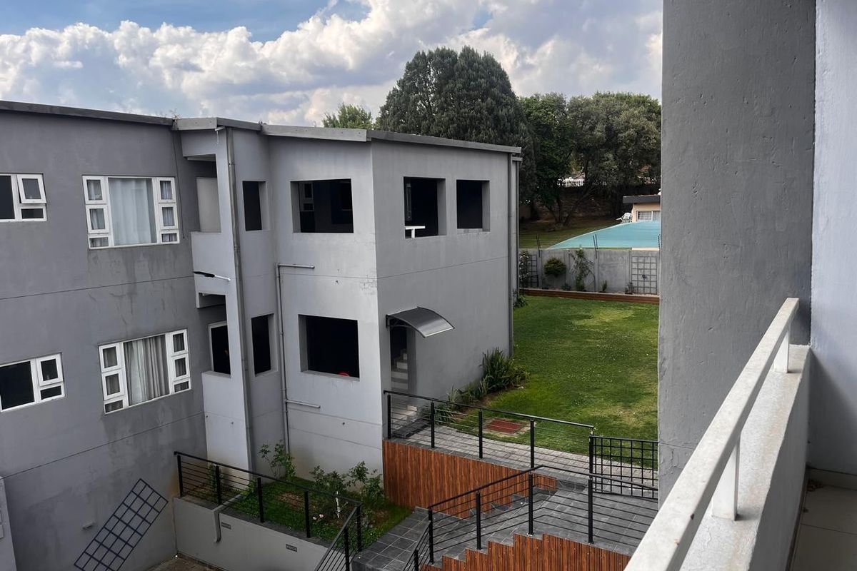 View from a balcony overlooking the rear of the apartment complex, featuring a landscaped garden, paved walkways, and a communal swimming pool area.
