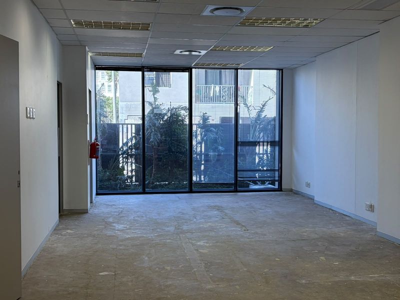 An empty, open-plan office space featuring large floor-to-ceiling windows, a suspended ceiling with recessed lighting, and an unfinished floor surface.