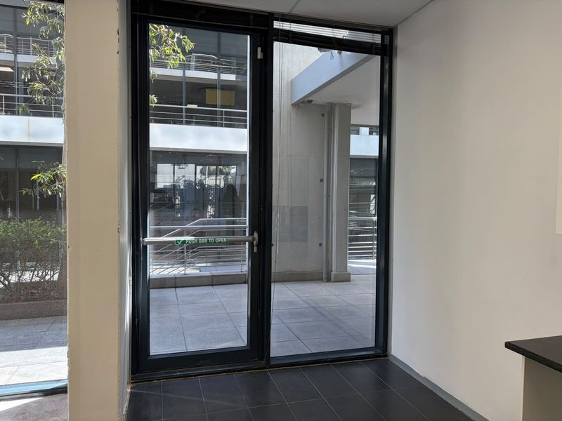 A clean, modern office entry featuring a glass door with a panic bar, dark tiled flooring, and a view of the exterior courtyard.