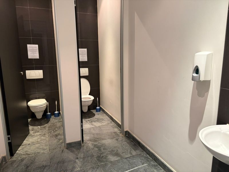 A clean, modern office restroom featuring two private toilet stalls with wall-mounted fixtures and a shared washbasin area with tiled finishes.