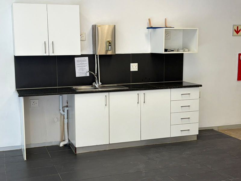 A functional office kitchenette featuring white cabinetry, a granite countertop, stainless steel sink, and a modern black tiled backsplash.
