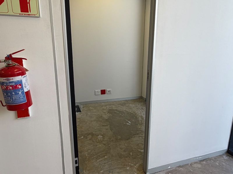 A small, empty office space featuring unfinished concrete flooring, electrical power points, and a mounted fire extinguisher in the adjacent hallway.