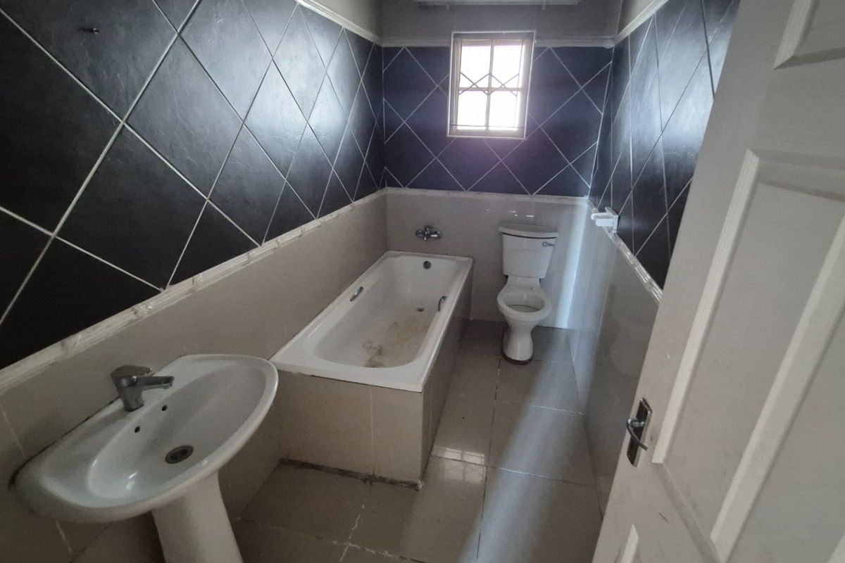 A dated bathroom featuring a pedestal basin, built-in bathtub, and toilet, finished with dark diamond-patterned wall tiles and neutral floor tiling.