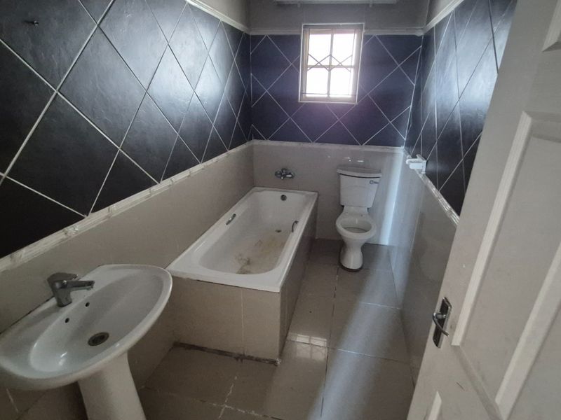 A dated bathroom featuring a pedestal basin, built-in bathtub, and toilet, finished with dark diamond-patterned wall tiles and neutral floor tiling.