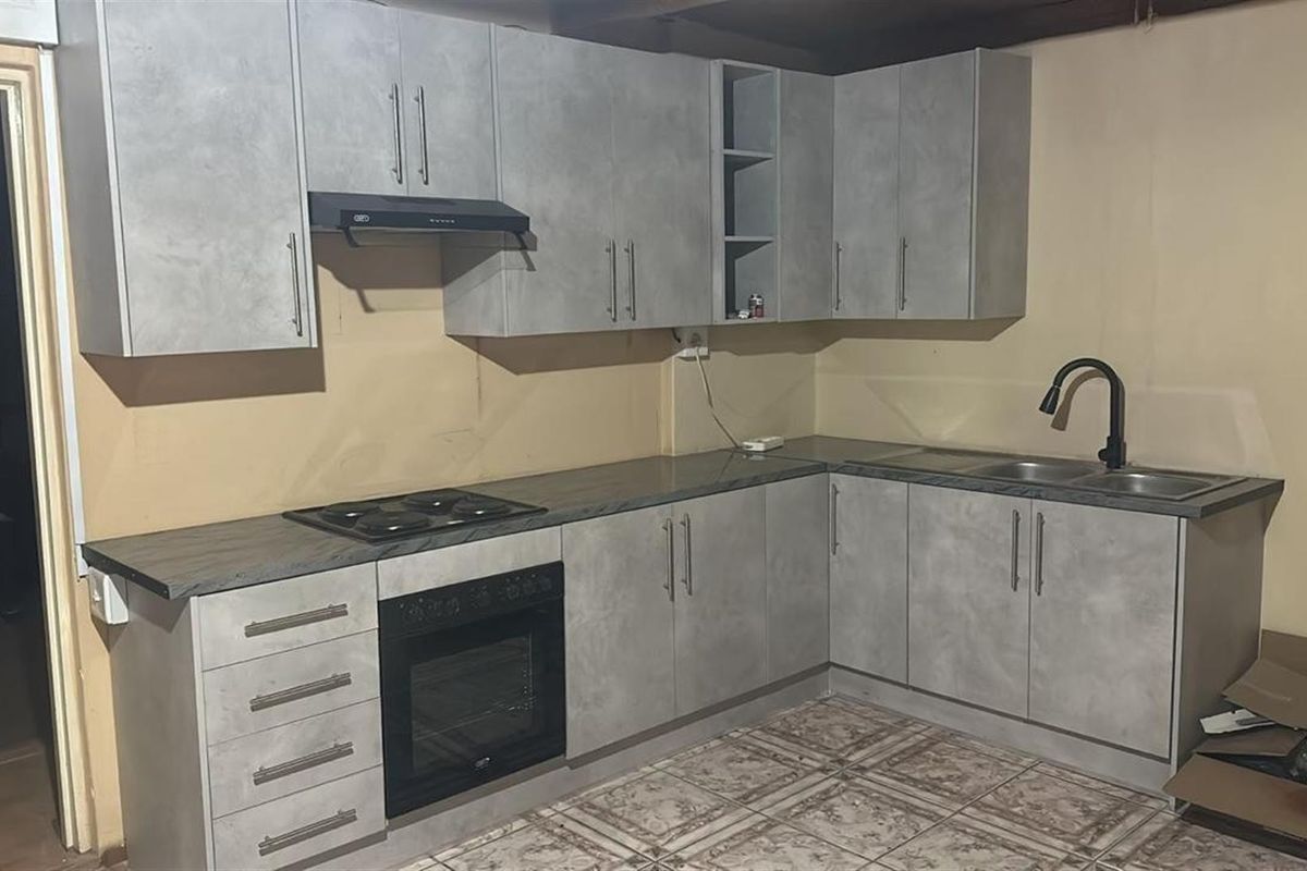 A functional kitchen featuring grey cabinetry, built-in oven and hob, and a double sink, currently requiring finishing touches to the wall surfaces.
