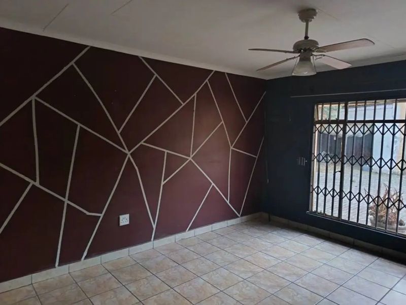 An empty living room featuring tiled flooring, a ceiling fan, and a decorative geometric accent wall with a security gate leading outdoors.