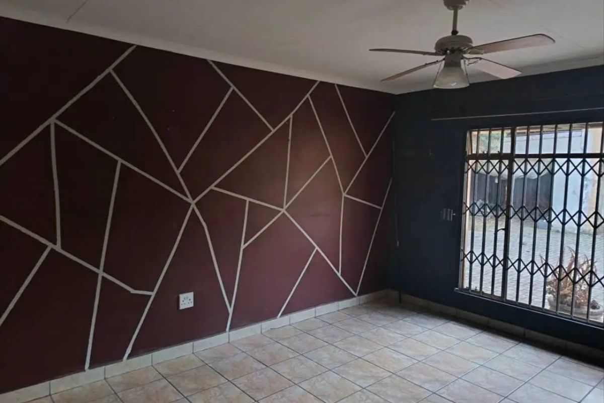 An empty living room featuring tiled flooring, a ceiling fan, and a decorative geometric accent wall with a security gate leading outdoors.