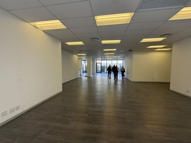 A spacious, empty open-plan office area featuring modern laminate flooring, a suspended ceiling with recessed lighting, and large windows providing natural light.