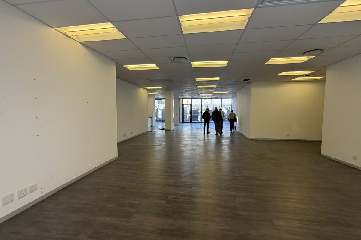 A spacious, empty open-plan office area featuring modern laminate flooring, a suspended ceiling with recessed lighting, and large windows providing natural light.