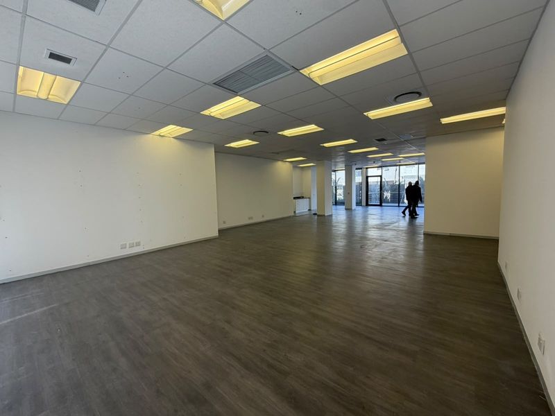 A spacious, empty open-plan office area featuring modern laminate flooring, a suspended ceiling with recessed lighting, and large glass doors leading to the exterior.
