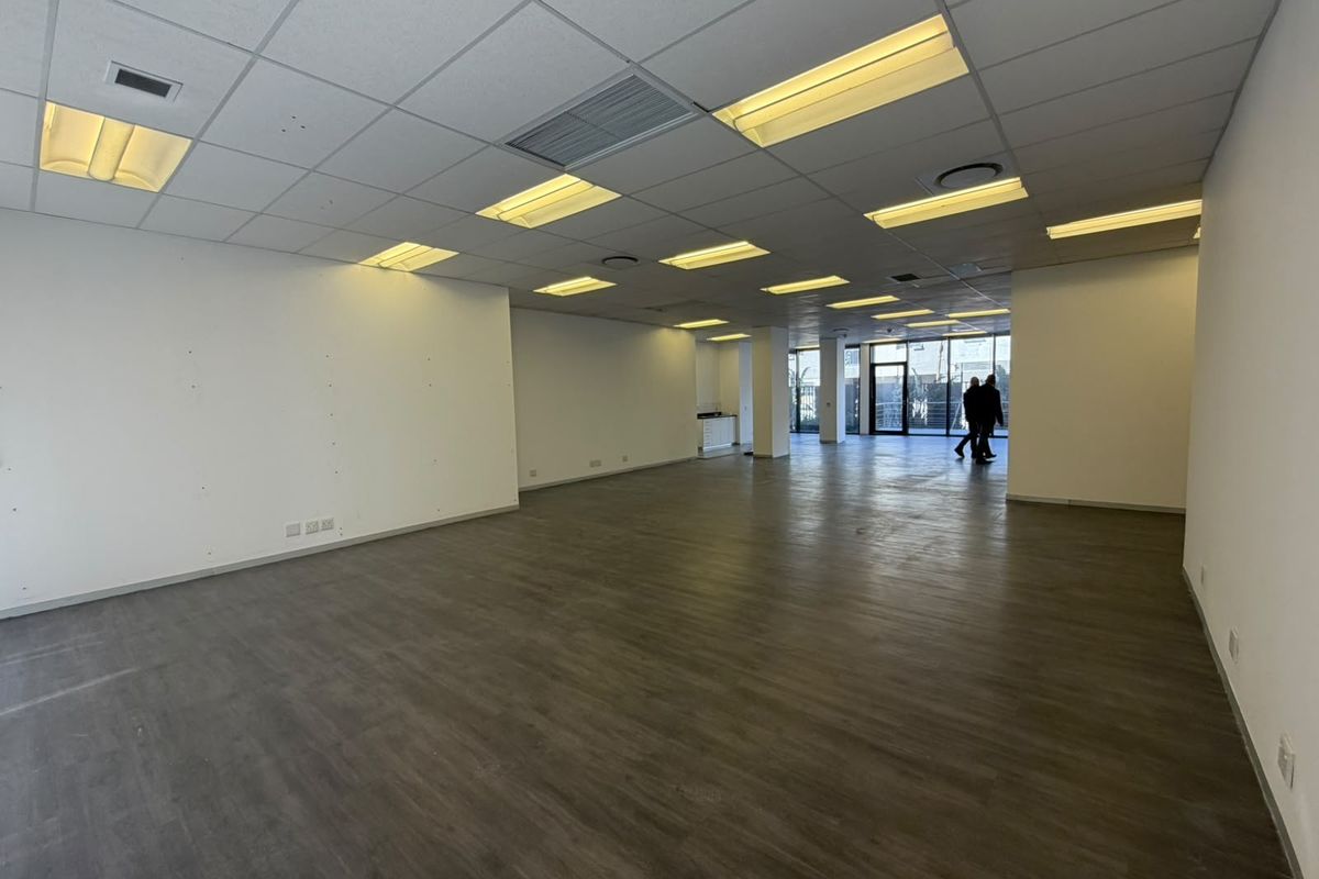A spacious, empty open-plan office area featuring modern laminate flooring, a suspended ceiling with recessed lighting, and large glass doors leading to the exterior.