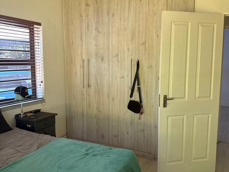 A bedroom featuring light wood-look built-in cupboards, laminate flooring, and a window with venetian blinds.