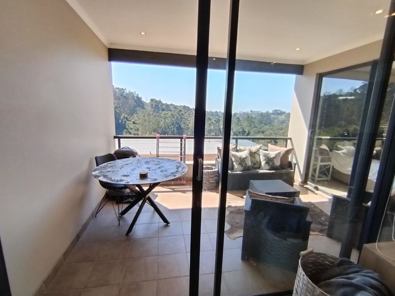 A covered balcony featuring tiled flooring, recessed lighting, and a glass balustrade, offering scenic views of the surrounding landscape.