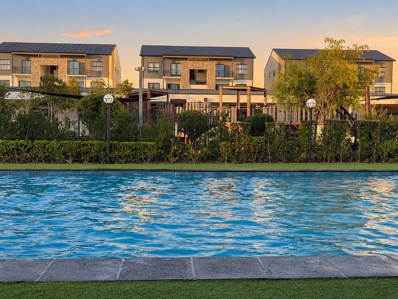 A well-maintained communal swimming pool area featuring stone coping, artificial turf, and perimeter fencing, set against a backdrop of modern apartment buildings.